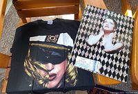 Photo showing Madonna tour T-shirt with a detailed portrait and the concert booklet with ticket stub on top.