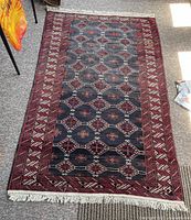 Full view of rectangular dark-colored Baluch hand knotted rug showing geometric diamond patterns and fringed edges on floor.