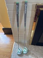 Two left-handed TaylorMade Kalea putters standing vertically leaning on a wall showing full shafts, grips, and heads with covers on the heads