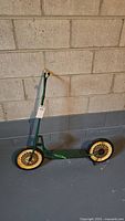Full view of a vintage green child's scooter with yellow spoked wheels against a concrete block wall.