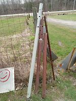 Photo of three metal jack posts leaning against wire fencing outdoors, including two rusty brown ones and one white one with multiple height adjustment holes.