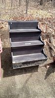 Full view of black molded plastic 4 step staircase standing on dirt ground outdoors with visible dirt and wear marks.
