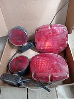 Four trailer lights in a cardboard box, including two square red lights and two round red lights with black housing, all with wiring attached.