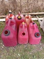 Photo showing six small fuel/gas cans in red and orange plastic with some dirt and wear, arranged outdoors.