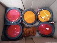 Seven trailer lights in a cardboard box showing three red round lights, two amber round lights, and two small rectangular amber lights.