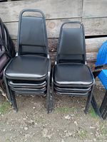 Photo showing two stacks of black chairs with metal frames. Chairs have padded black seats and backs, stacked outdoors on dirt ground.