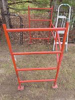 Red metal folding scaffolding frame standing on grass next to a ladder and other items, showing front view and base with feet plates.