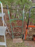 Green metal dolly cart with two vertical handles and a flat base plate, positioned outdoors against foliage.