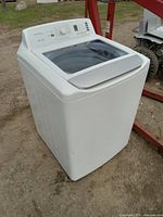 Full view of white Insignia top load washing machine standing outdoors on dirt ground.