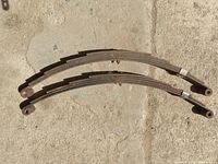 Top view of two curved multi-leaf trailer leaf springs placed on concrete, showing oval eyelets with metal bushings at each end.