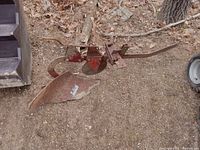 Photo of a single metal plough implement sitting on dirt ground next to a tire and stairs, showing the blade and attachment points.
