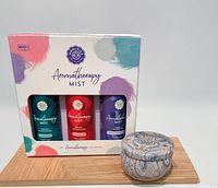 Photo of boxed set of three Aromatherapy Mist bottles with colorful packaging alongside a small patterned tin candle container on a wooden surface.
