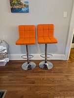 Two bright orange adjustable bar stools with-square stitched cushioned seats and backrests, chrome pedestal bases and footrests shown against a gray wall and hardwood floor.