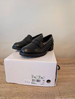 Pair of black women's chunky heel loafer shoes on top of their original white box. Visible stud detailing on heel collar and chunky tread sole. Box label shows size 7 wide.