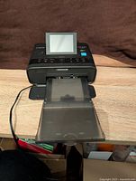 Front view of the Canon Selphy CP1300 photo printer on wooden surface, screen raised, and paper tray extended, with power cord attached.