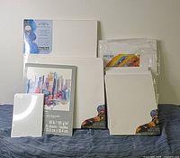 Various canvas panels and watercolor pad arranged on cloth surface. Includes large and small canvases, 32-pack canvas panels in plastic wrapping, and watercolor pad with city skyline image on cover.