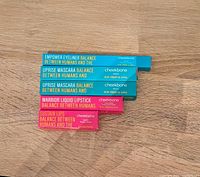 Stacked side view of five boxed cosmetic items from Cheekbone Beauty: Empower Eyeliner (blue box), two Uprise Mascaras (blue boxes), Warrior Liquid Lipstick and Sustain Lips Lipstick (pink boxes) on wooden surface.