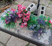 Image showing a bunch of assorted artificial flowers and greenery laying on a glass-top outdoor table with visible dirt and leaves from outdoor exposure.