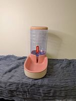 Single image showing the plastic gravity water bowl in beige and pink with clear water reservoir and beige lid, placed on a dark cloth surface against a plain wall.