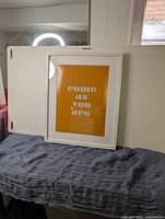 IKEA white RIBBA frame with orange print reading 'come as you are', placed on a blue cloth surface in front of a white wall and a window.