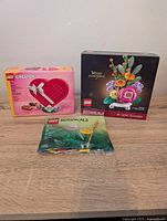 Three Lego sets displayed including one boxed Lego Creator Heart Box, one boxed Lego Botanicals Mixed Flowerpot set, and one sealed Lego Botanicals Field Flowers bag.