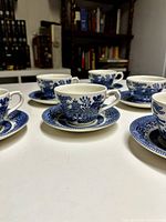 Close-up of six Blue Willow tea cups with matching saucers arranged on a white table, showing classic blue and white pattern details