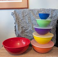Six nesting bowls in rainbow colors stacked on each other with lids on four bowls. Largest red and smallest blue bowls without lids. One bowl placed separately on left side.