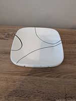 Top view of a single square Corelle plate showing white surface with black curved line design.