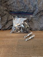 A plastic ziplock bag filled with stainless steel clothes pins along with several laid out in front on a wooden surface.