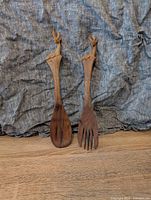 Pair of hand-carved wooden salad tongs featuring detailed animal heads and a spoon and fork design, laid flat on fabric background