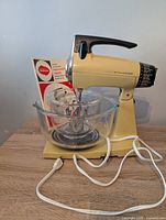 Vintage Sunbeam Mixmaster stand mixer in yellow with black handle on a table with two glass mixing bowls and instruction manual behind it.