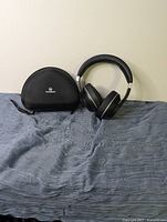 Black wireless over-ear Vankyo headphones, Vankyo half-moon shaped carrying case on a grey fabric surface against a plain light wall.