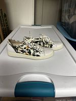 Side view of white women's sneakers with floral print on upper, white toe cap and sole, placed on a plastic surface.