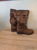 Side and angled view of the pair of brown boots showing straps, buckles, knit cuff, zipper and lugged sole