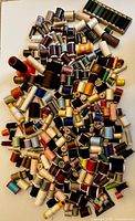 Over 150 mixed unopened and lightly used thread spools in various neutral and some colored tones scattered on a white surface.