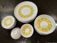 Top view of the complete set arranged on a dark surface showing 8 dinner plates, 8 side plates, 8 large bowls, and 8 small bowls with a yellow floral wreath design on white porcelain.