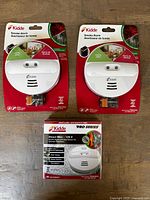 Top view of two Kidde battery-powered smoke alarms (Pi9010CA) and one direct wire Kidde smoke alarm (Pi2010CA) all unopened in original packaging on wooden surface.