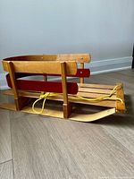 Side view of vintage wooden sleigh showing natural wood and red side rails with attached yellow pull rope.