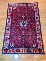 Full view of Persian rug showing red field, cream floral border, and fringe on wood floor.