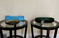 Three Ikebana containers displayed on two round glass-top pedestal tables against a plain wall.