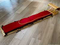 Full side view of wooden toboggan sled with padded red seat and yellow rope trim laid on wood floor.