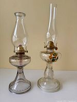 Two vintage glass oil lamps shown side by side on a white surface against a light beige background. One clear glass lamp with floral decoration, one smoky tinted glass lamp.
