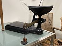 Side view of the black metal vintage scale showing the large bowl pan, balancing beam, and hanging weight on a glass table.