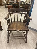 Front view of an antique spindle back wooden chair showing the curved armrests, spindle back, and contoured seat on a hardwood floor.