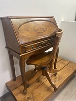 Side angle view of antique wooden desk with carved oval design on drop-front panel and swivel chair on caster wheels.