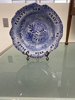 Blue and white ceramic decorative plate displayed on a black metal stand showing inscription and leaf/flower design.