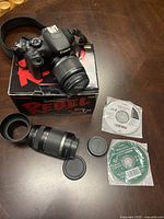 Camera body on original Rebel T2i box with telephoto lens, lens hood, lens caps and software CDs displayed on wood surface