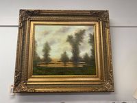 Front view of the framed oil painting showing the landscape scene with trees and water under a cloudy sky within an ornate gold frame.