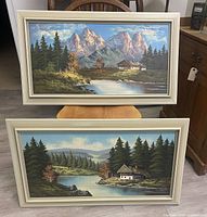 Front view of two framed oil paintings showing mountain and lakeside landscapes, standing on a chair.