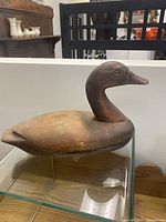 Side angle view of brown wooden duck decoy on glass surface showing its profile and head shape.